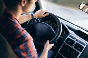 Person driving a car on a sunny day, hands on the steering wheel.